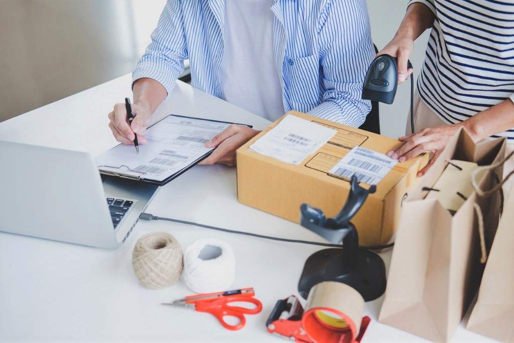 3D, concept image to represent reverse logistics, a man and a woman in stripey shirts, scanning a barcode on a parcel, and checking the corresponding code on a clipboard, whilst also using a laptop, preparing an item for return logistics.